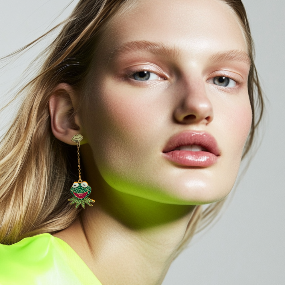 Close-up of a person wearing a bright yellow shirt and yellow gold plated Kermit the frog drop earrings with green crystals by Couture Kingdom against a neutral background
