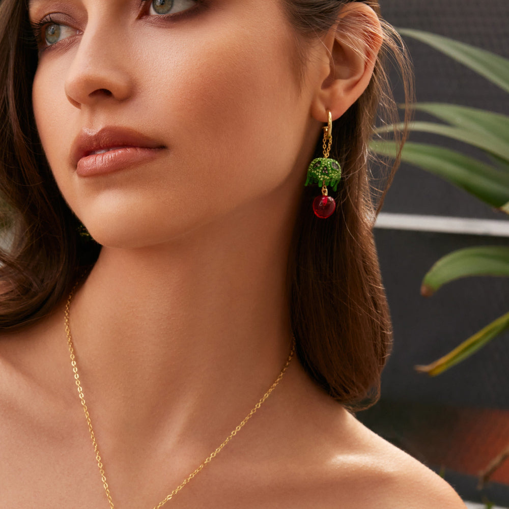 Yellow gold plated Snow White Poison Apple Drop Earrings with mini hoop and green crystal poison apple hanging from hoop by Couture Kingdom worn on model with green leafy background