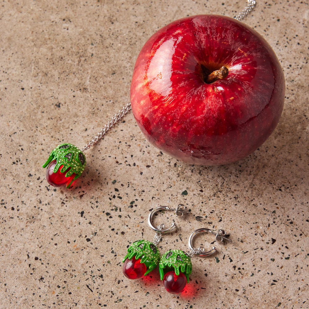 White Gold Plated Snow White Poison Apple Drop Earrings with mini hoop and matching necklace by Couture Kingdom flat lay near red apple on a brown marble background